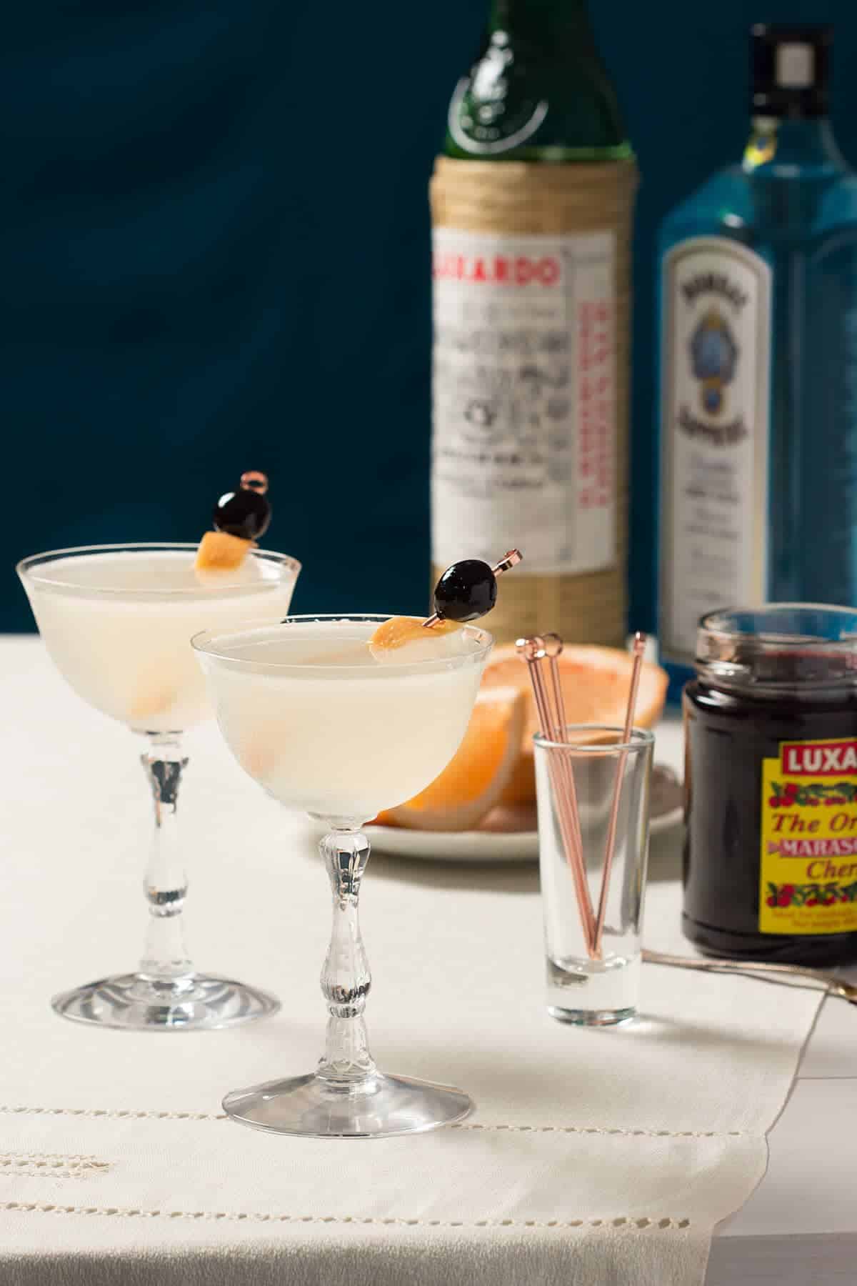 Two seventh heaven cocktails next to bottles of liquor and a jar of maraschino cherries.