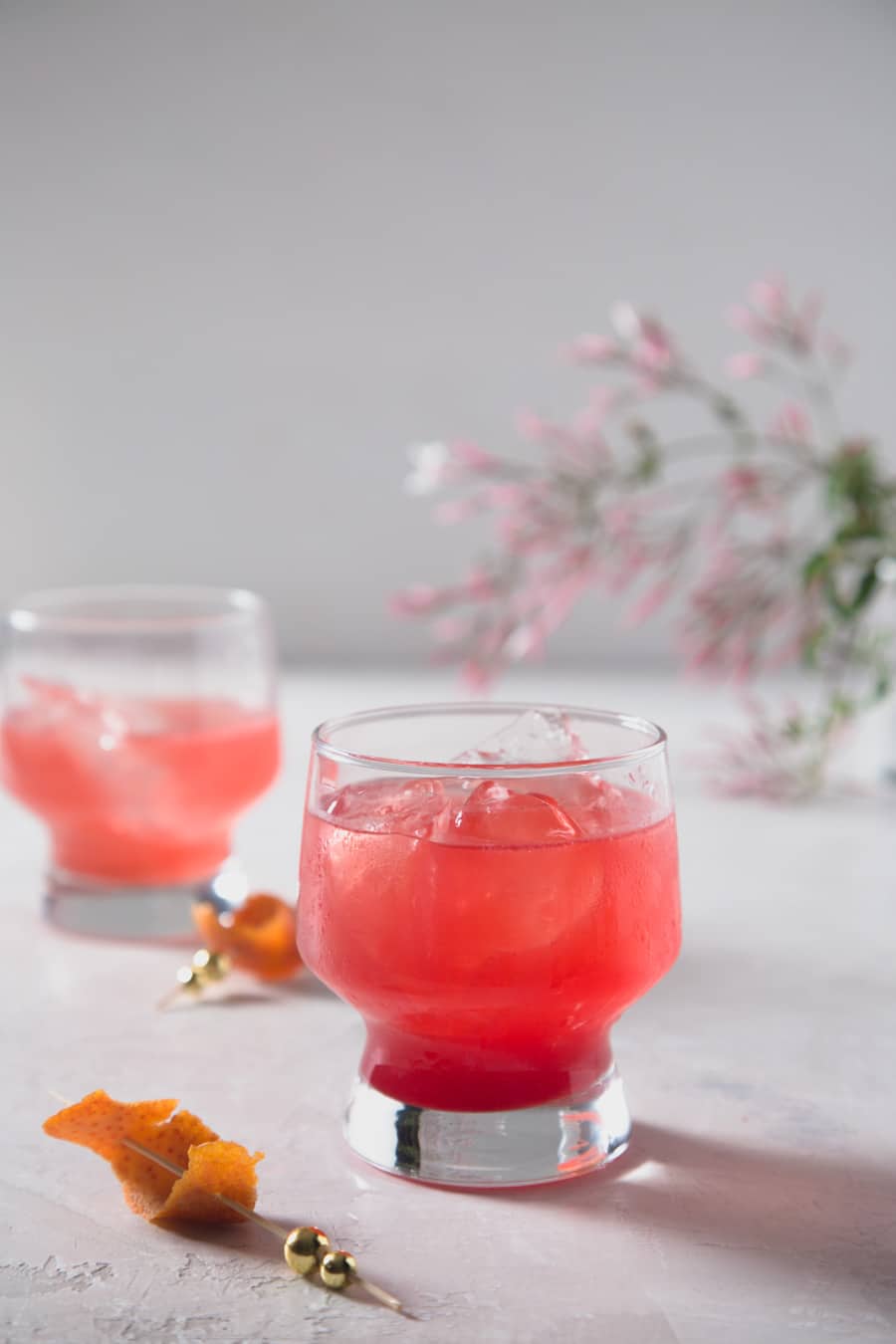 Partially drunk blood orange gin and tonic cocktail with a orange twist garnish on the surface in the foreground and jasmine flowers in background.