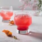 Close up of a partially drunk blood orange gin and tonic cocktail with a orange twist garnish on the surface in the foreground and jasmine flowers in background.