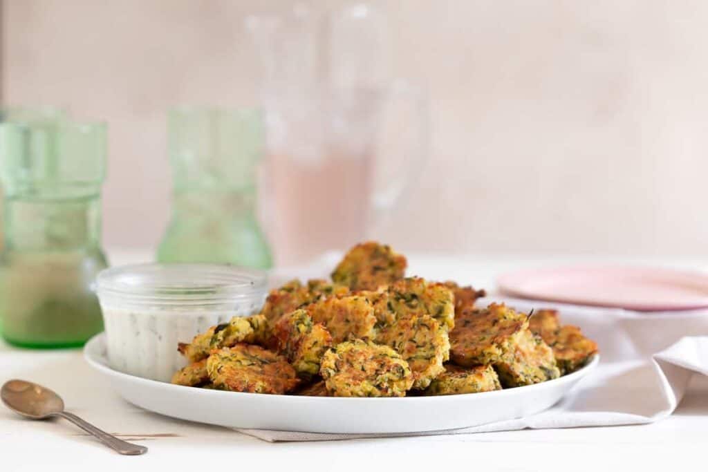 Platter of baked zucchini and cheddar fritters with a jar of dipping sauce and drinks in the background.