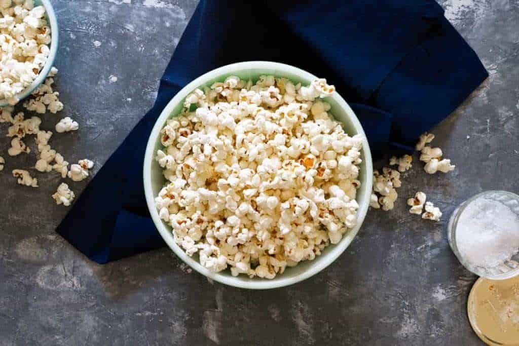 horizontal image of a bowl of popcorn on a dark blue napkin with popcorn on the surface