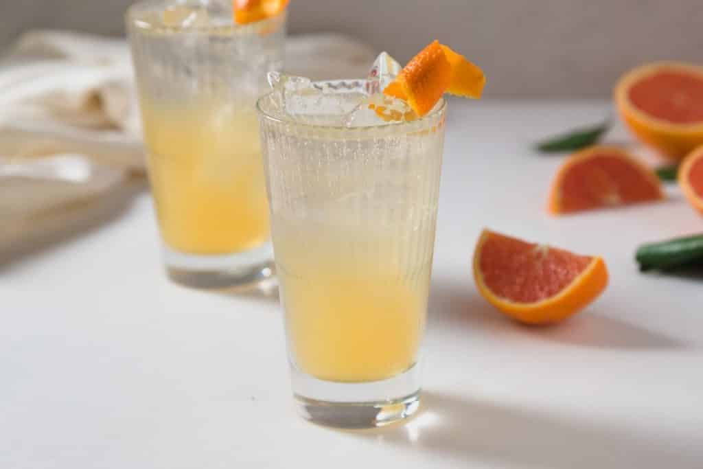 two orange colored cocktails with an orange twist as a garnish and wedgest of oranges in the background.