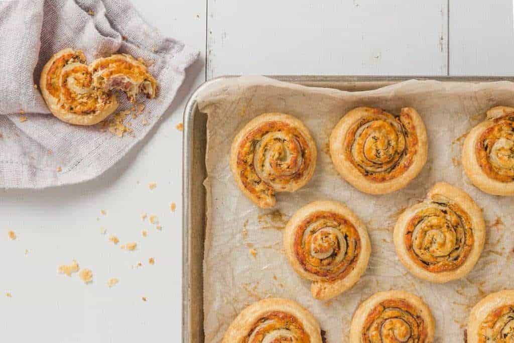 parchment line baking sheet with baked white cheddar prosciutto pinwheels and a broken pinwheel on a napkin