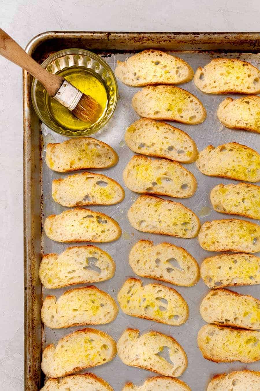 Using a pastry brush dipped in oil to brush oil onto sliced pieces of baguette.
