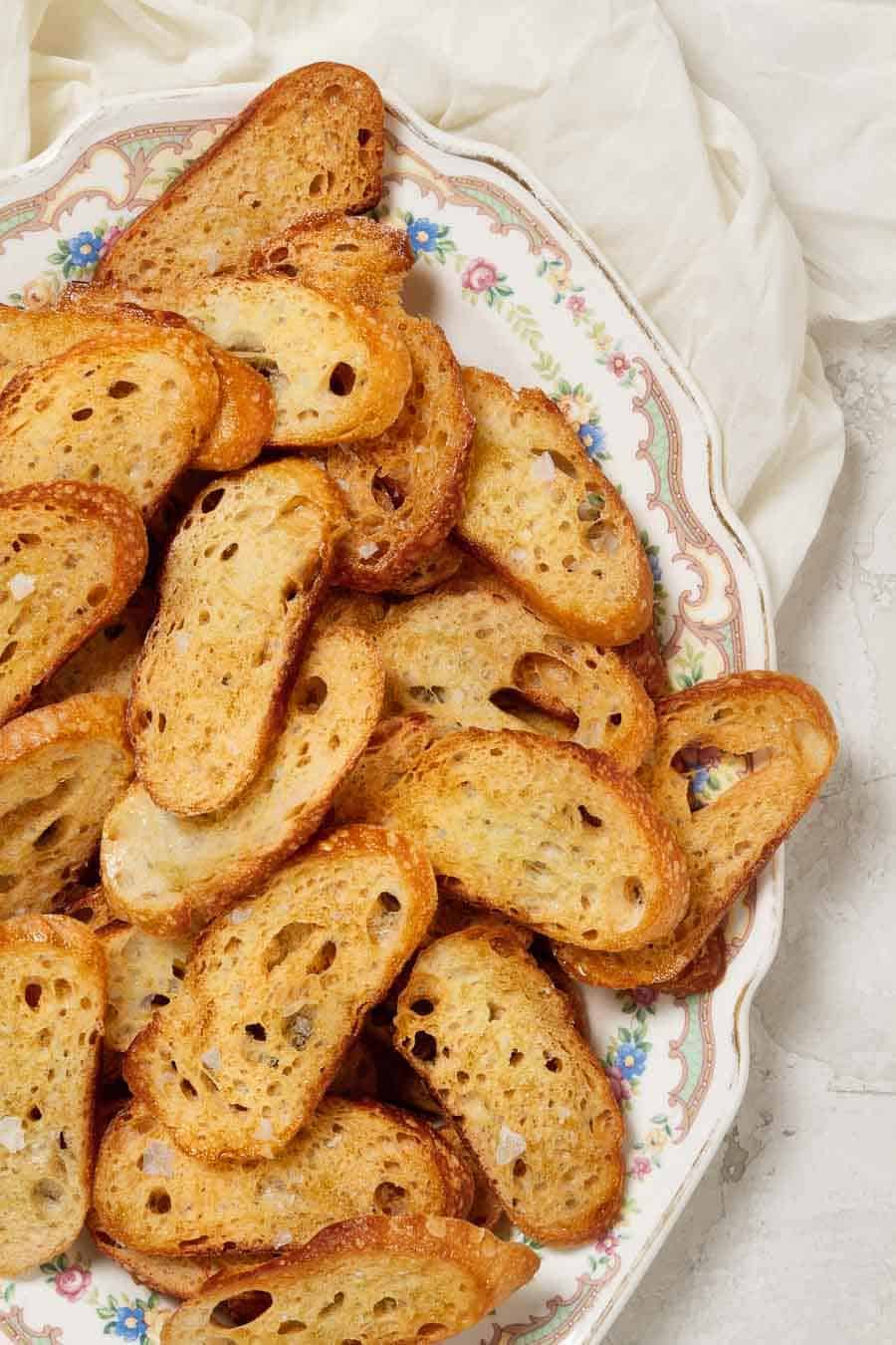 a close up of baked crostini, topped with flake salt, piled on an antique platter