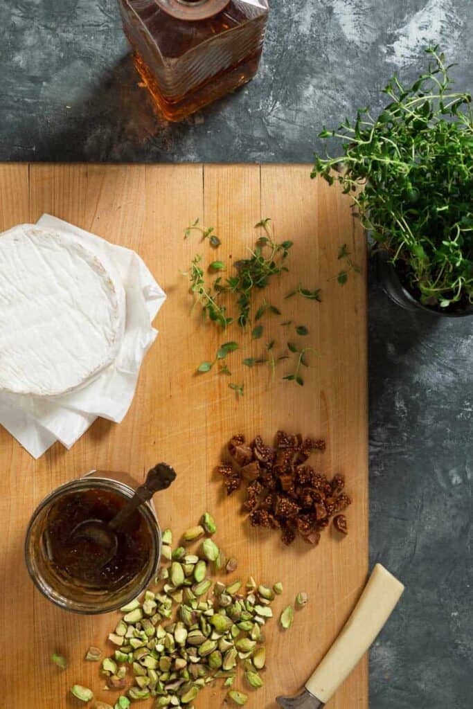 Chopped pistachios and dried figs, a jar of fig jam and thyme leaves are on a wooden cutting board with a decanter of brandy and a thyme plant next to it.