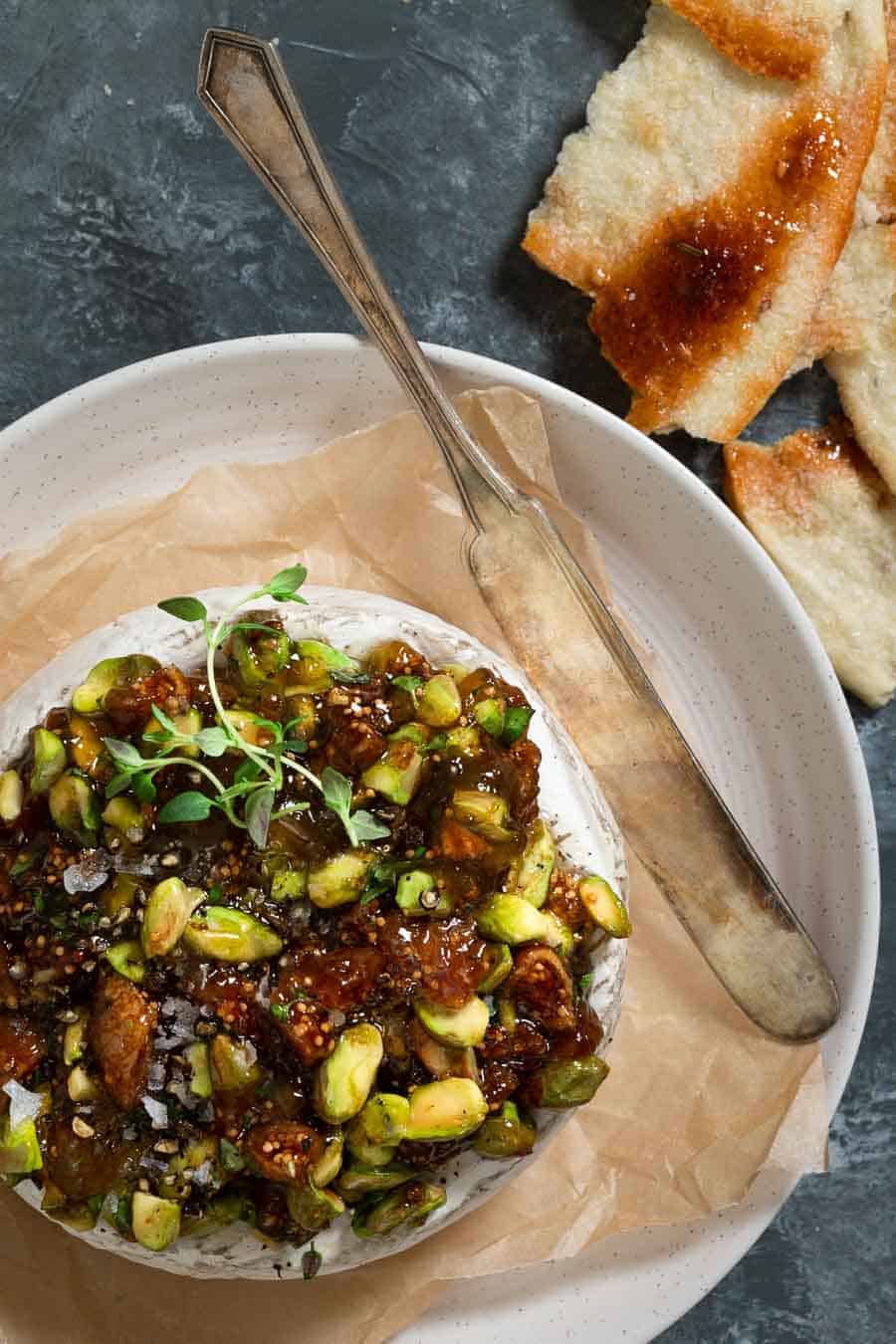 A wheel of baked brie topped with fig jam, chopped dried figs, pistachios and thyme of a plater ready to be cut into.