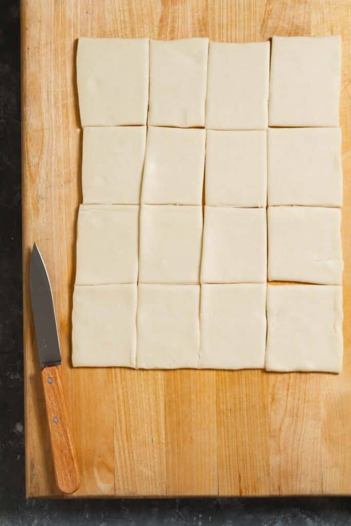 a sheet of puff pastry on a wooden cutting board cut into sixteen rectangles with a wooden handled pairing knife next to it.