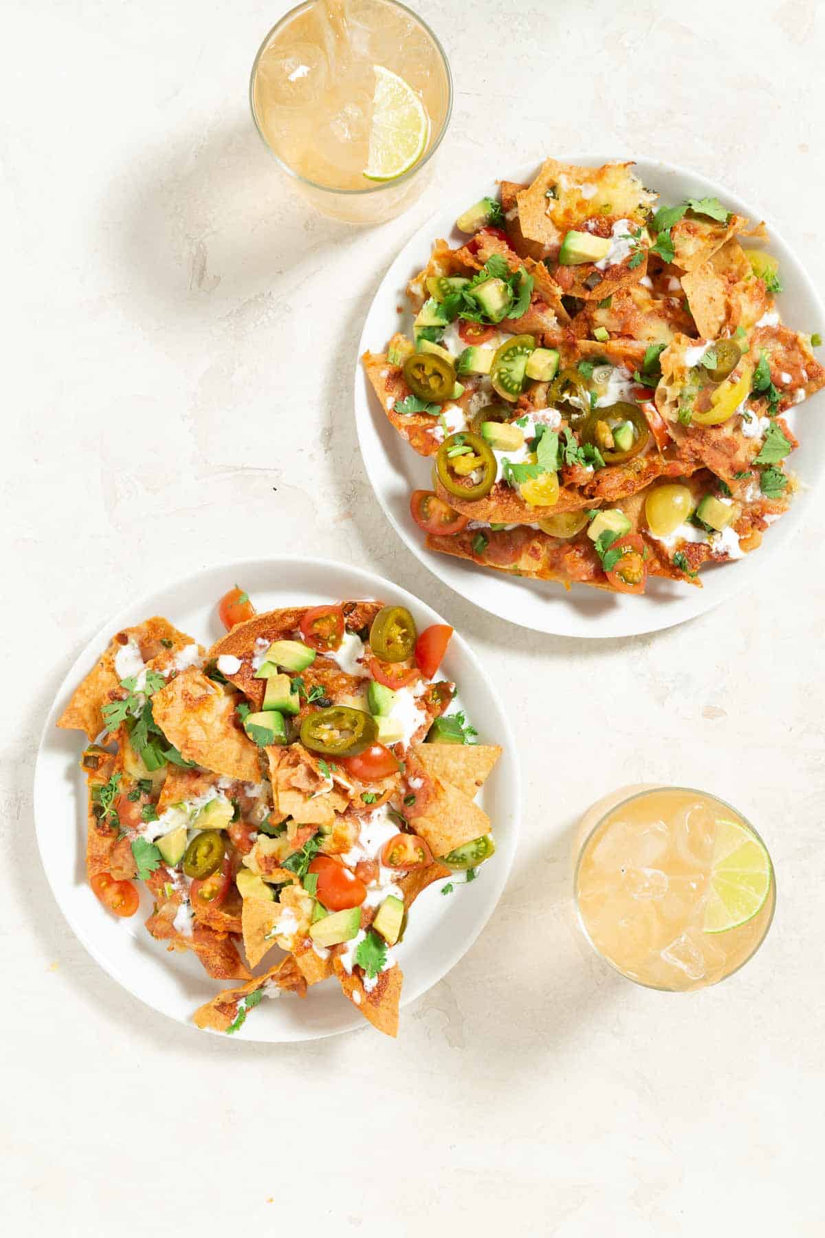 2 servings of vegetarian nachos on white plates. Paloma cocktails in glasses on the side.