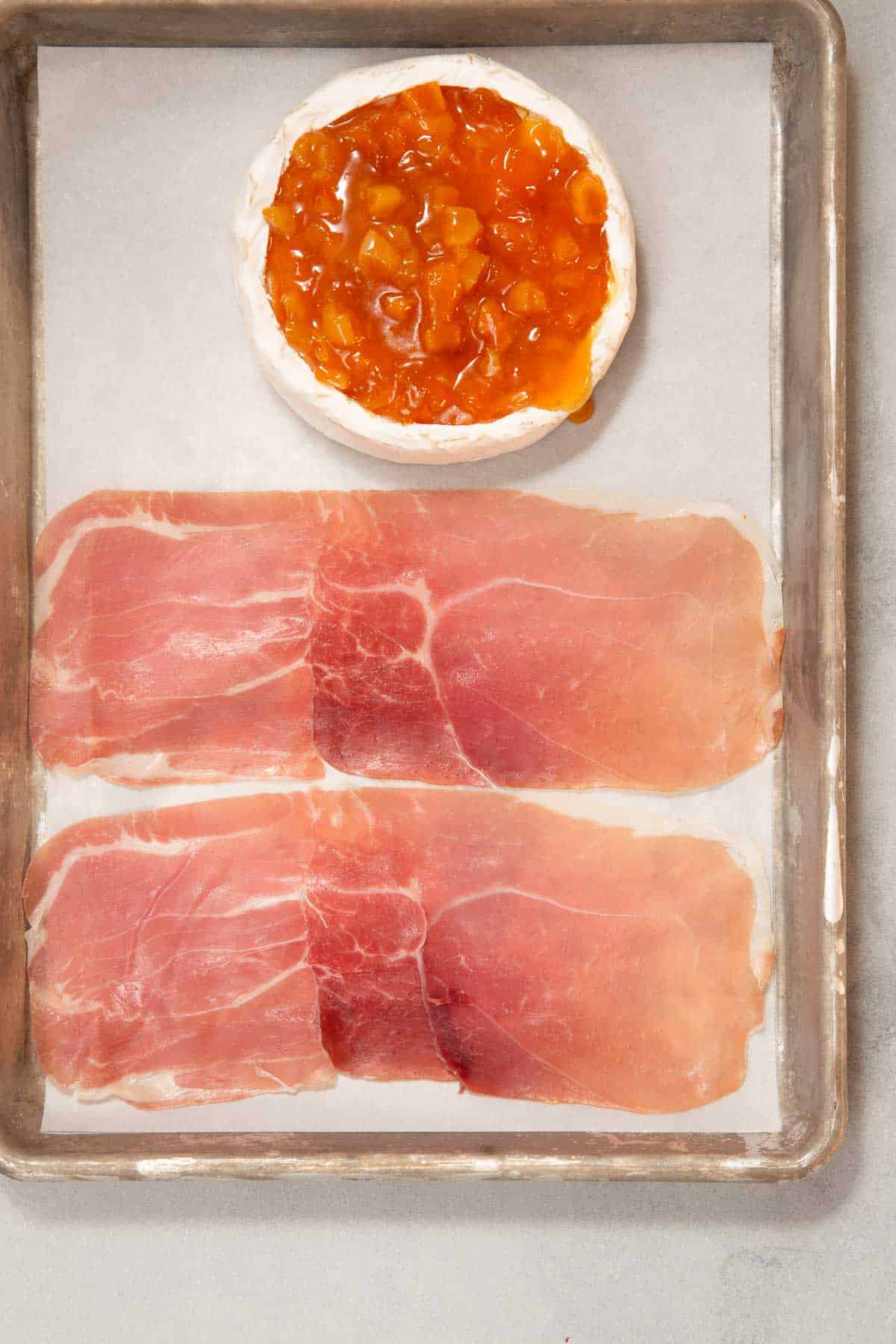 Wheel of brie and slices of raw deli meat on a baking sheet