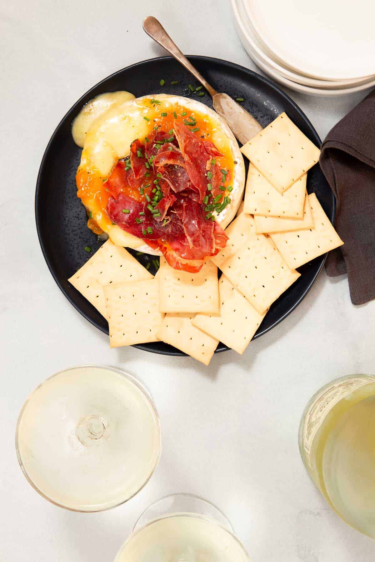 Black plate with a wheel of brie and meat on top. Crackers on the side.