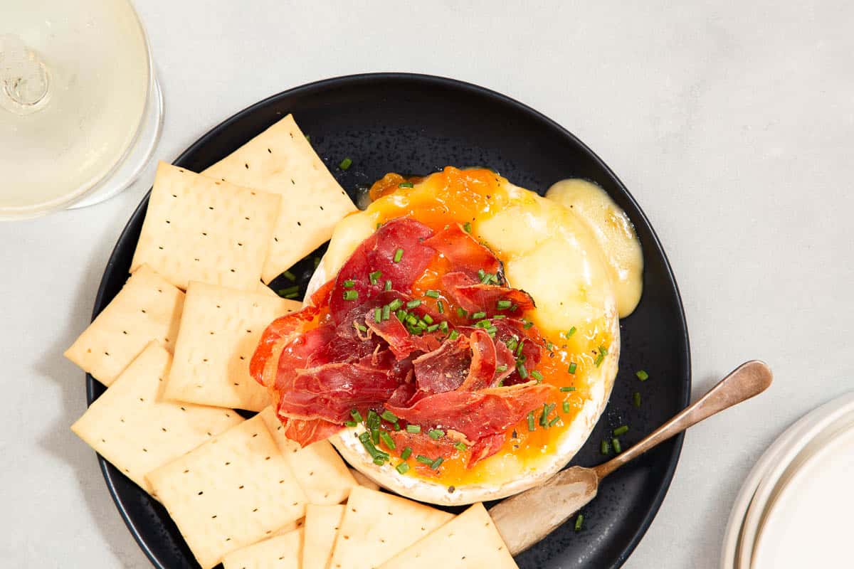 Close up of a wheel of brie on a black plate with meat and preserves on top with crackers on the side