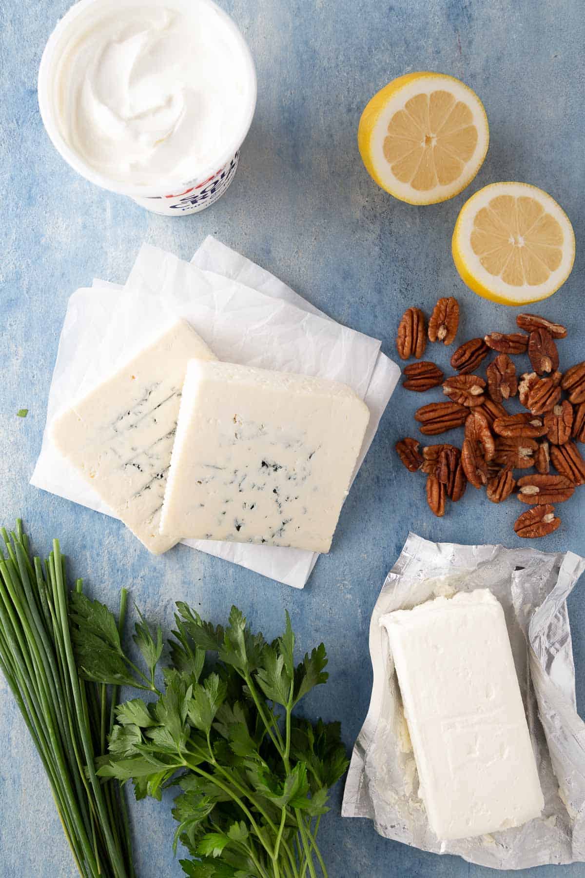 slices of blue cheese, cream cheese, sour cream, lemon, herbs, and pecans on a blue counter.