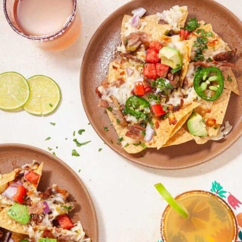 two brown plates holding carnitas nachos next to lime slices and drinks.