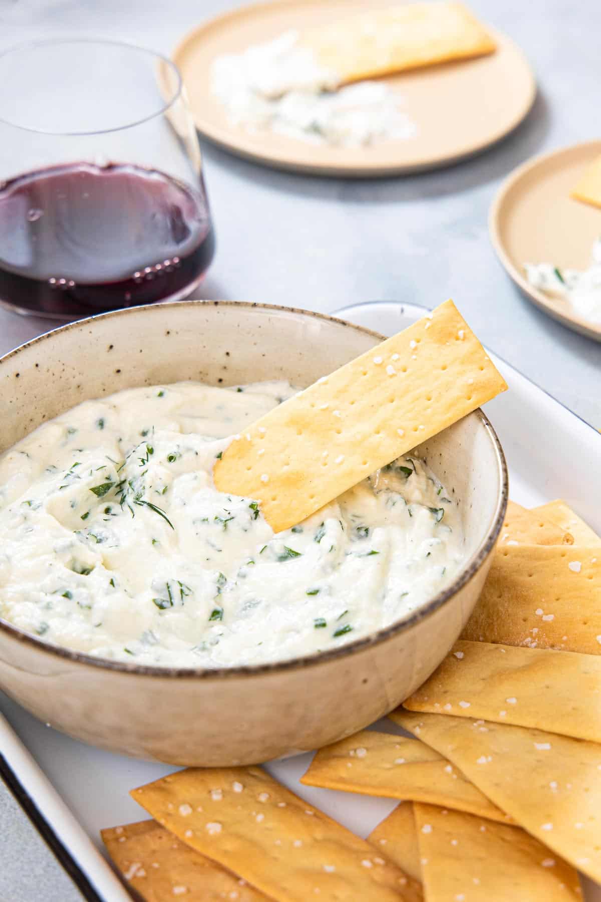 a bowl of herb ricotta dip surrounded by crackers and a glass of red wine.