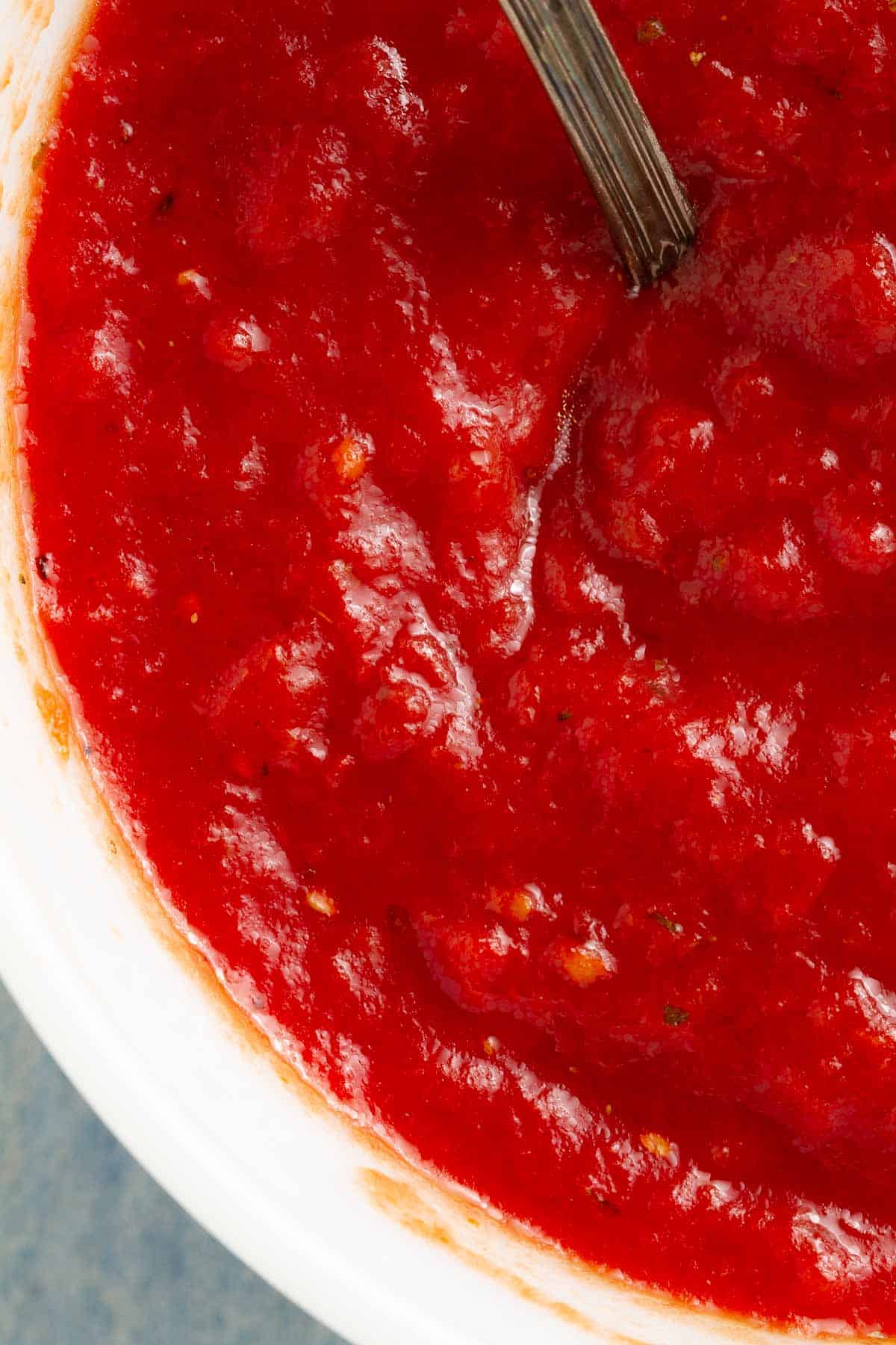 Close up of a white bowl filled with a red tomato sauce with a spoon handle coming out of it.