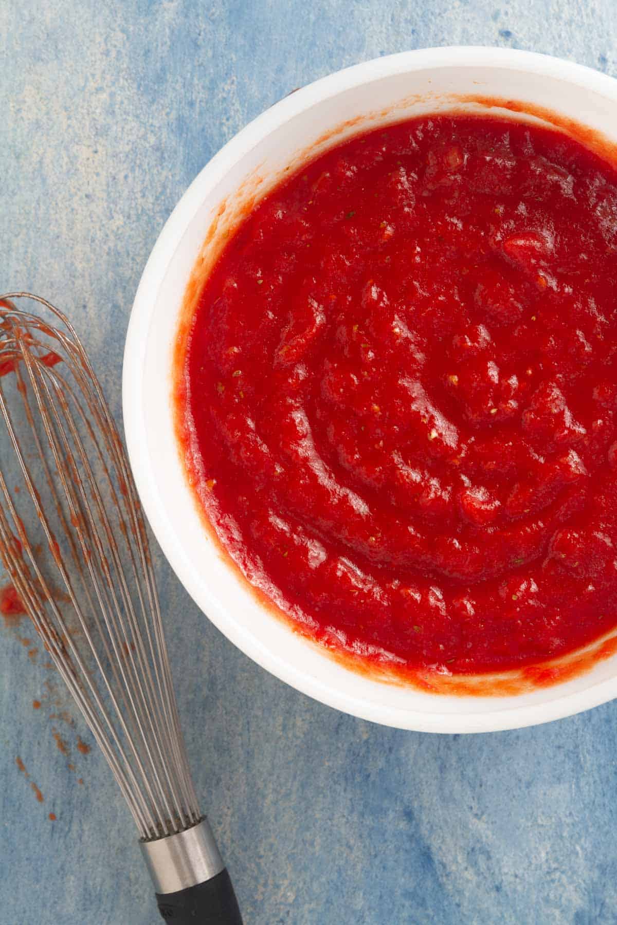 Overhead of a white bowl filled with a pizza sauce and a whisk next to it.