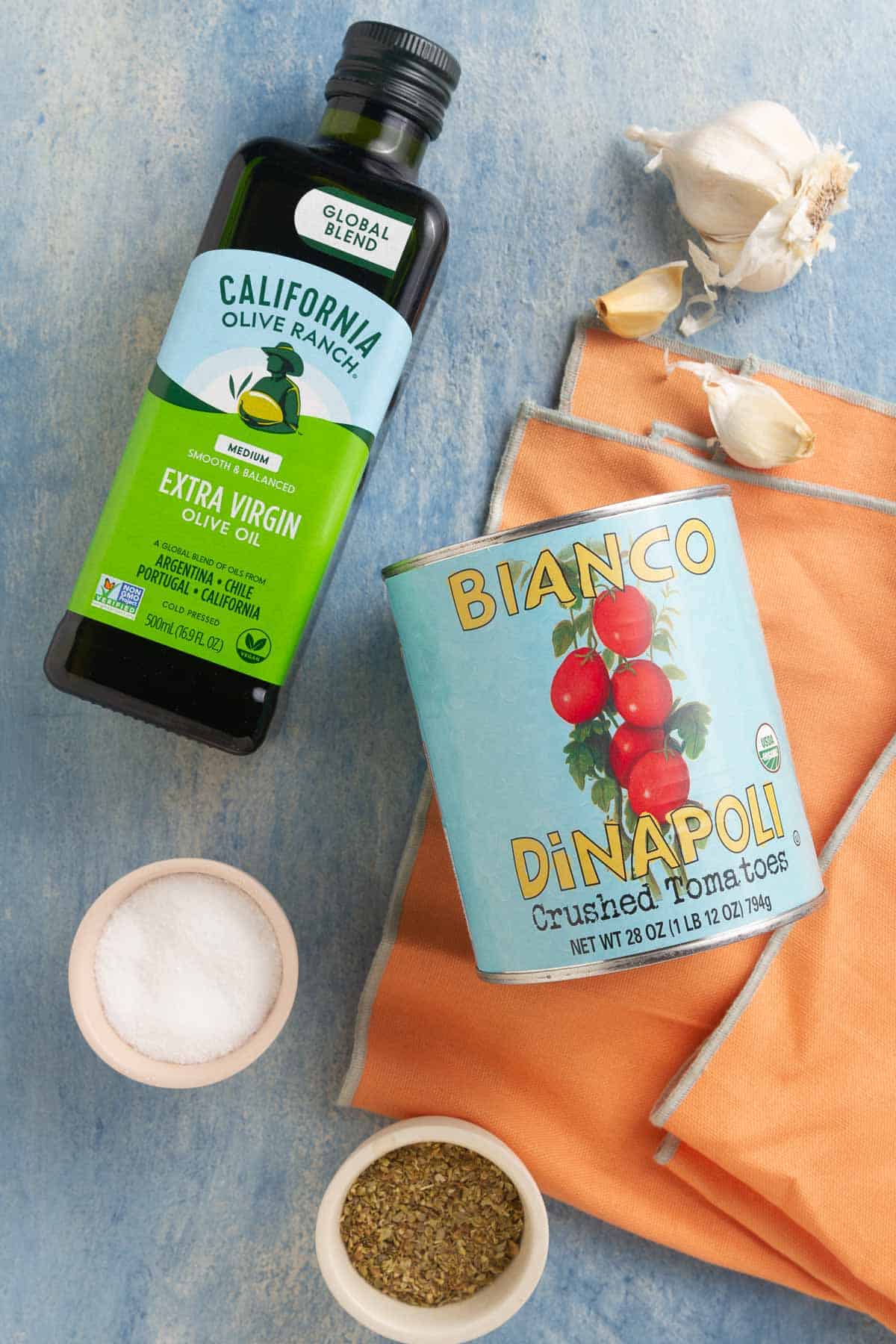 A bottle of olive oil, a head of garlic, a small bowl of salt and a small bowl of oregano, and a can of crushed tomatoes on a blue surface.