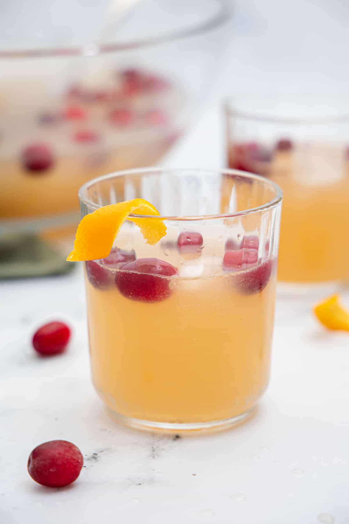 A small glass filled with a light orange beverage, floating cranberries and ice cubes with an orange twist garnish.