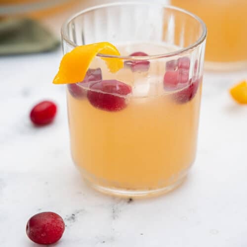 Close up of a small glass filled with an orange liquid, ice, cranberries and garnished with an orange twist.