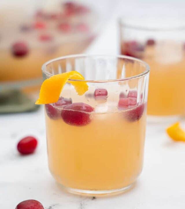 A small glass filled with a light orange beverage, floating cranberries and ice cubes with an orange twist garnish.
