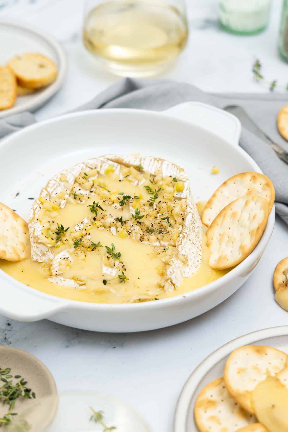 baked brie with garlic and thyme in a white baking dish with crackers inside the dish.