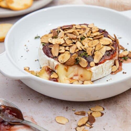 melted brie cheese topped with toasted almonds and jam in a white baking dish.