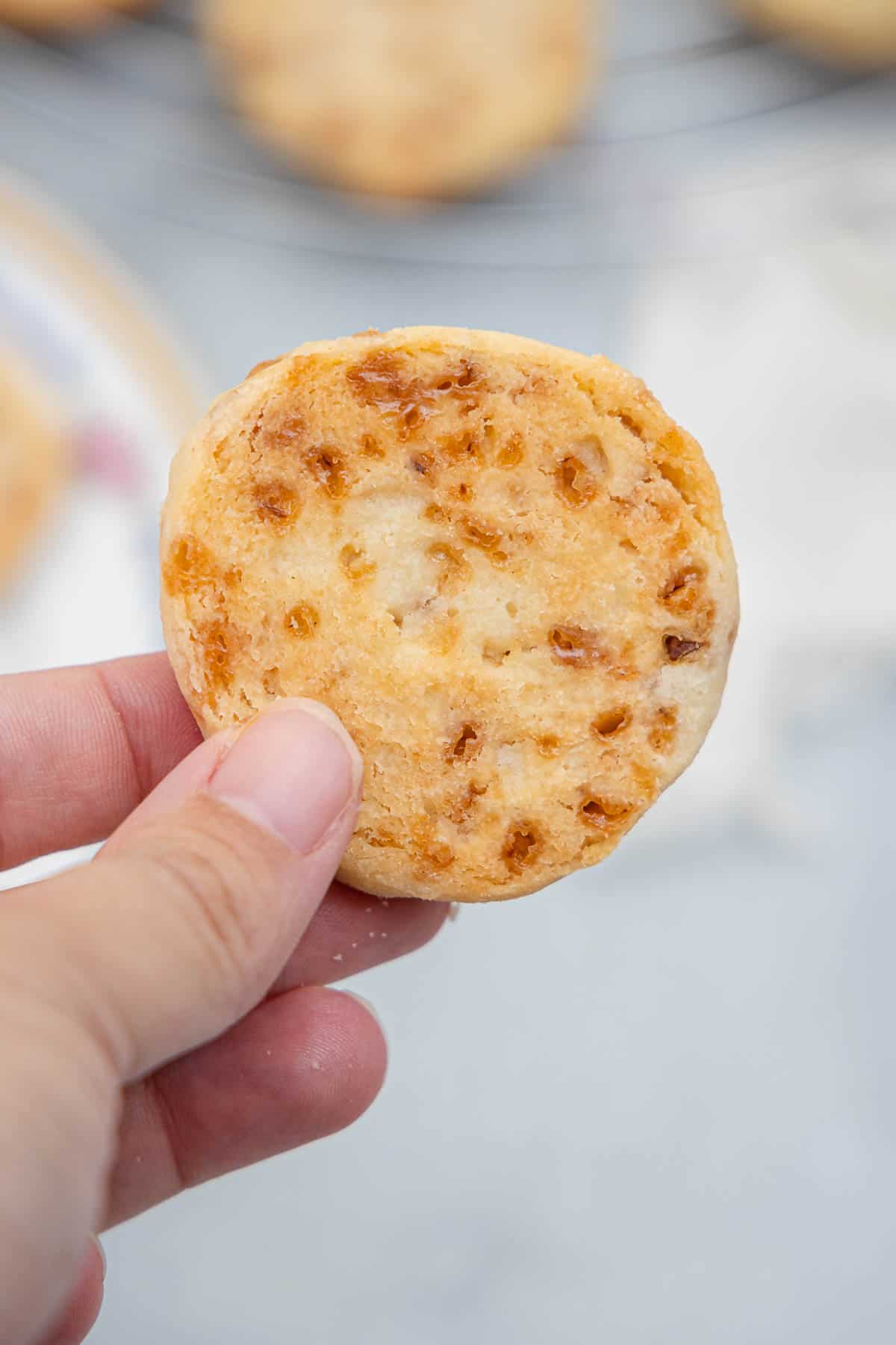 A hand holding a toffee shortbread cookie with the backside facing the camera so the doneness of the cookie can be shown.