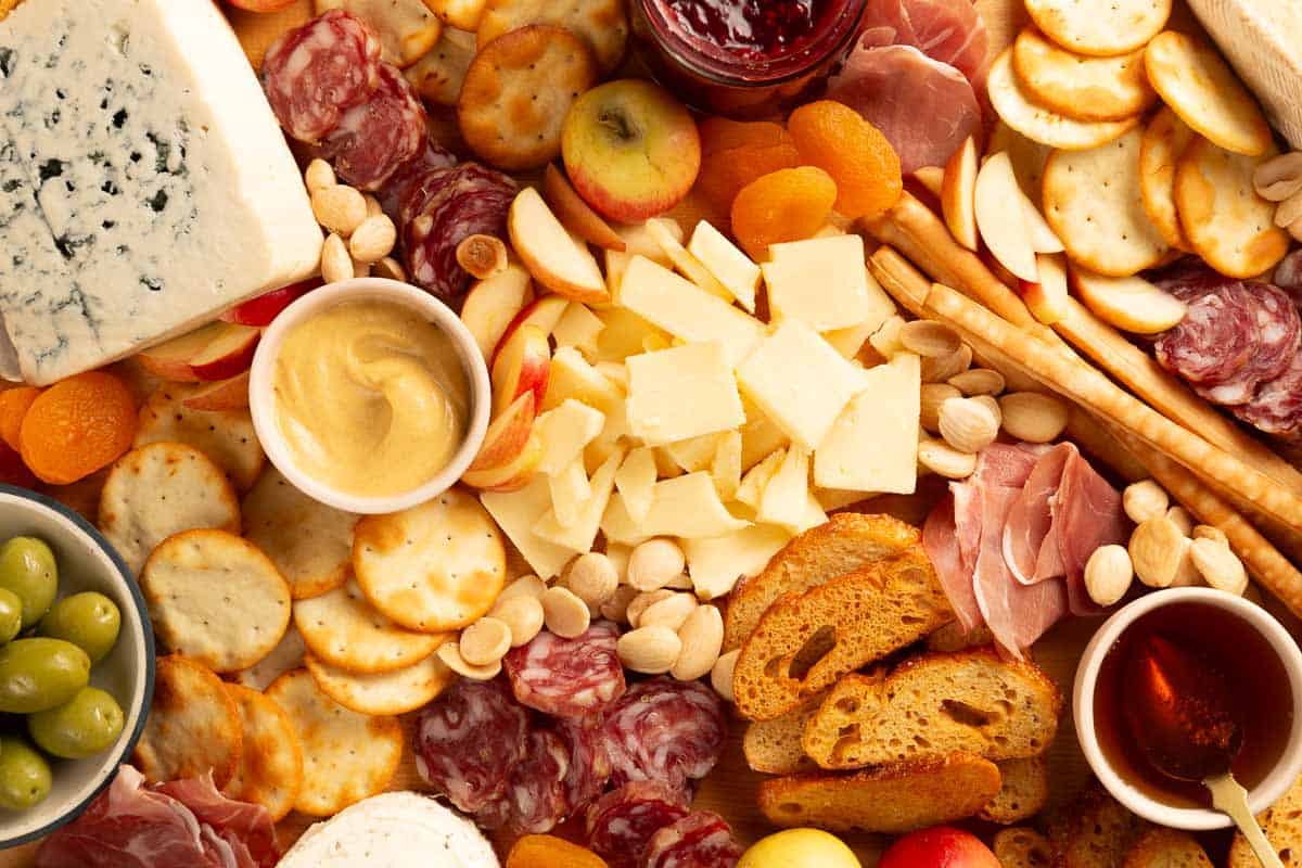Close up overhead of a wooden cutting board with cheeses, jars of condiments, crackers, olives, charcuterie, jam, honey and whole fruit on it.v