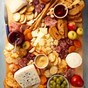 Overhead of a wooden cutting board with cheeses, jars of condiments, crackers, olives, charcuterie, jam, honey and whole fruit on it.