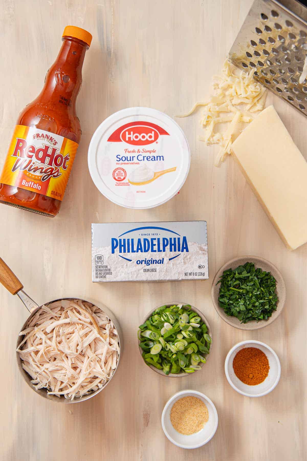 Overhead of a bottle of wing sauce, a container of sour cream, a cheese grater, a block of white cheese, small bowls individually filled with chopped parsley, green onions, spices, a measuring cup filled with shredded white meat chicken, and a block of cream cheese in the center.
