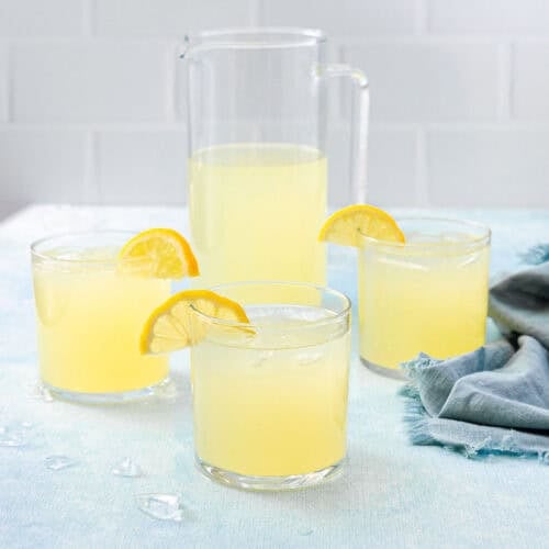 Three glasses filled with ice and a yellow liquid in them each with a lemon slice garnish and a pitcher of the same liquid in the background.
