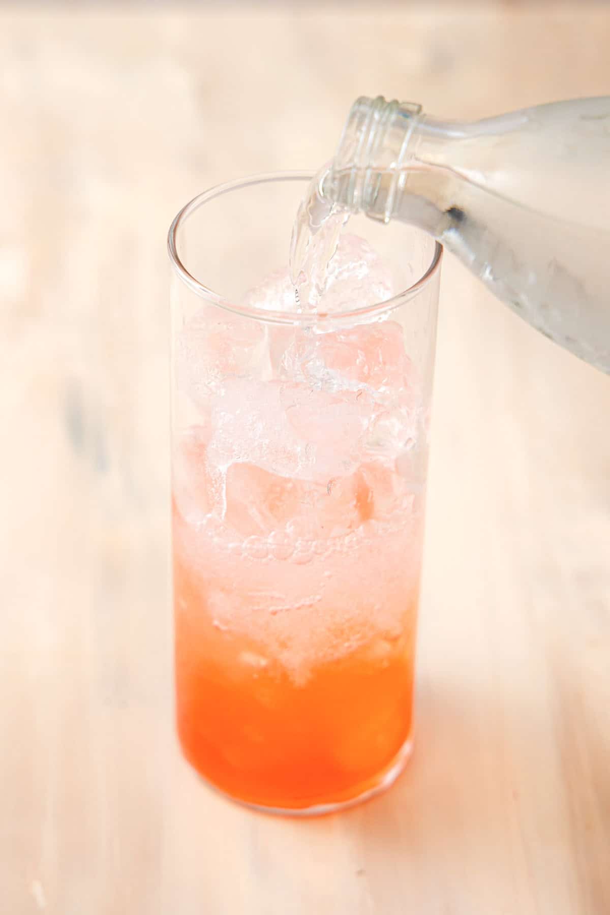 A tall glass filled with ice and a light pink liquid with sparkling mineral water being poured into it.