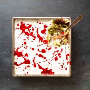 Overhead of a square metal pan with a top layer of sour cream and a hot sauce "blood splatter" with a spoon in the top right corner that has scooped out a serving of the dip.