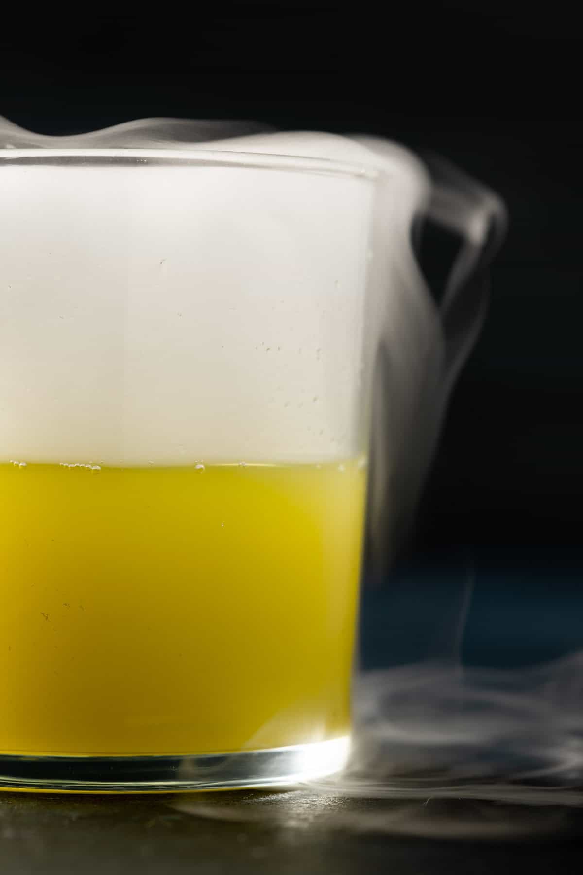 Extreme close up of a glass filled half way with a muddy green colored liquid and a white smoke filling the remaining half of the glass with smoke spilling out of the glass.