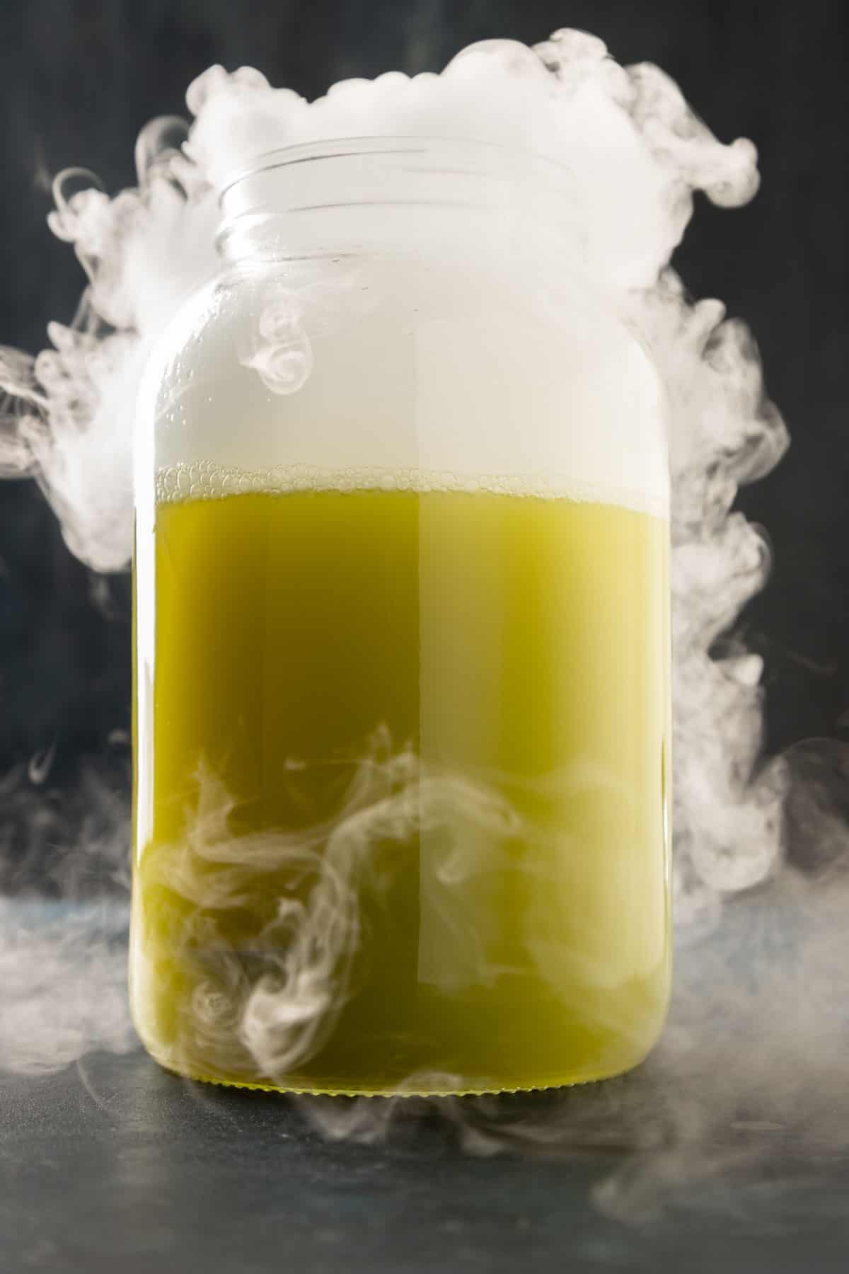 A large punch jar filled with a muddy green liquid and a white smoke billowing out of the top and around it.