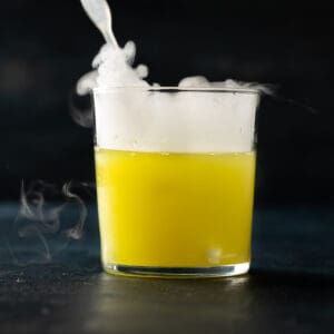 Close up of a glass filled half way with a muddy green colored liquid and a white smoke filling the remaining half of the glass with smoke spilling out of the glass.
