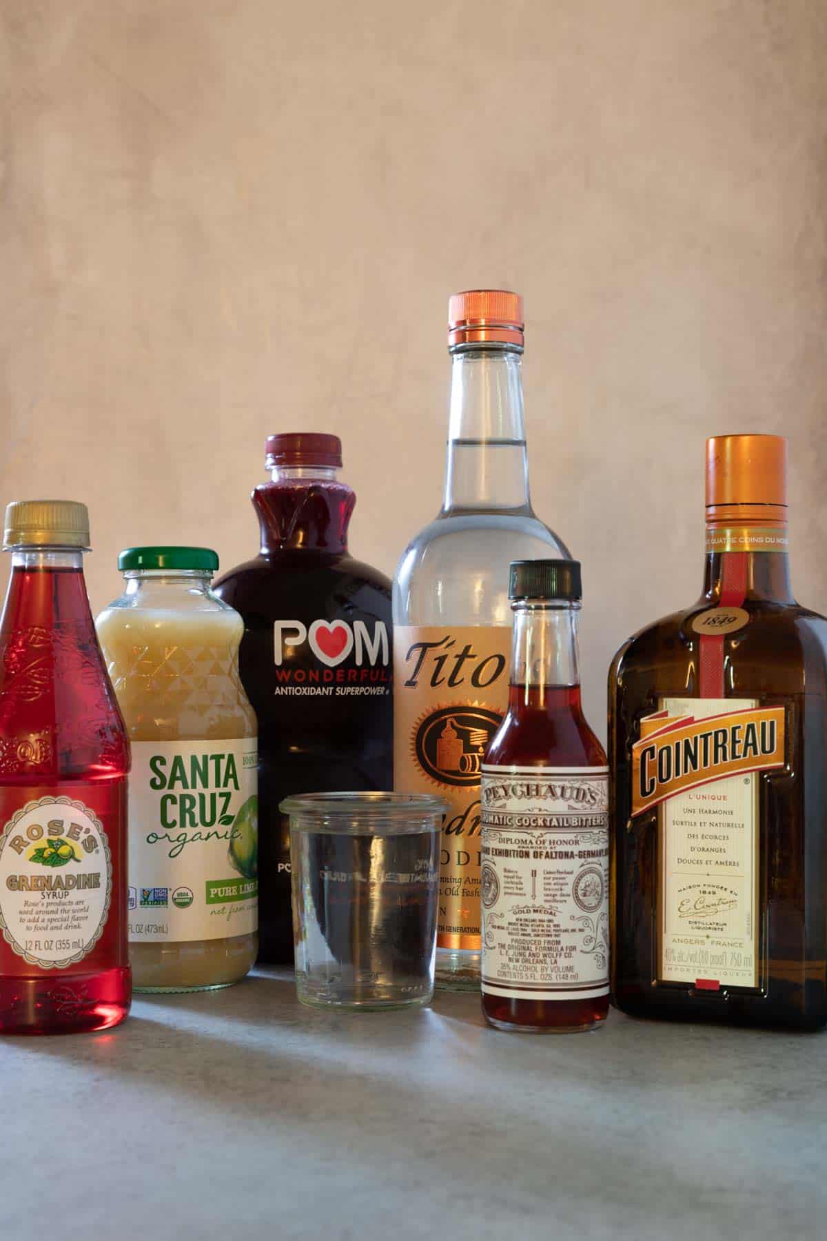 Ingredients to make the cocktail from left to right: bottle of grenadine, bottle of lime juice, bottle of pomegranate juice, a small jar of water, a bottle of vodka, a bottle of bitters and a bottle of cointreau.
