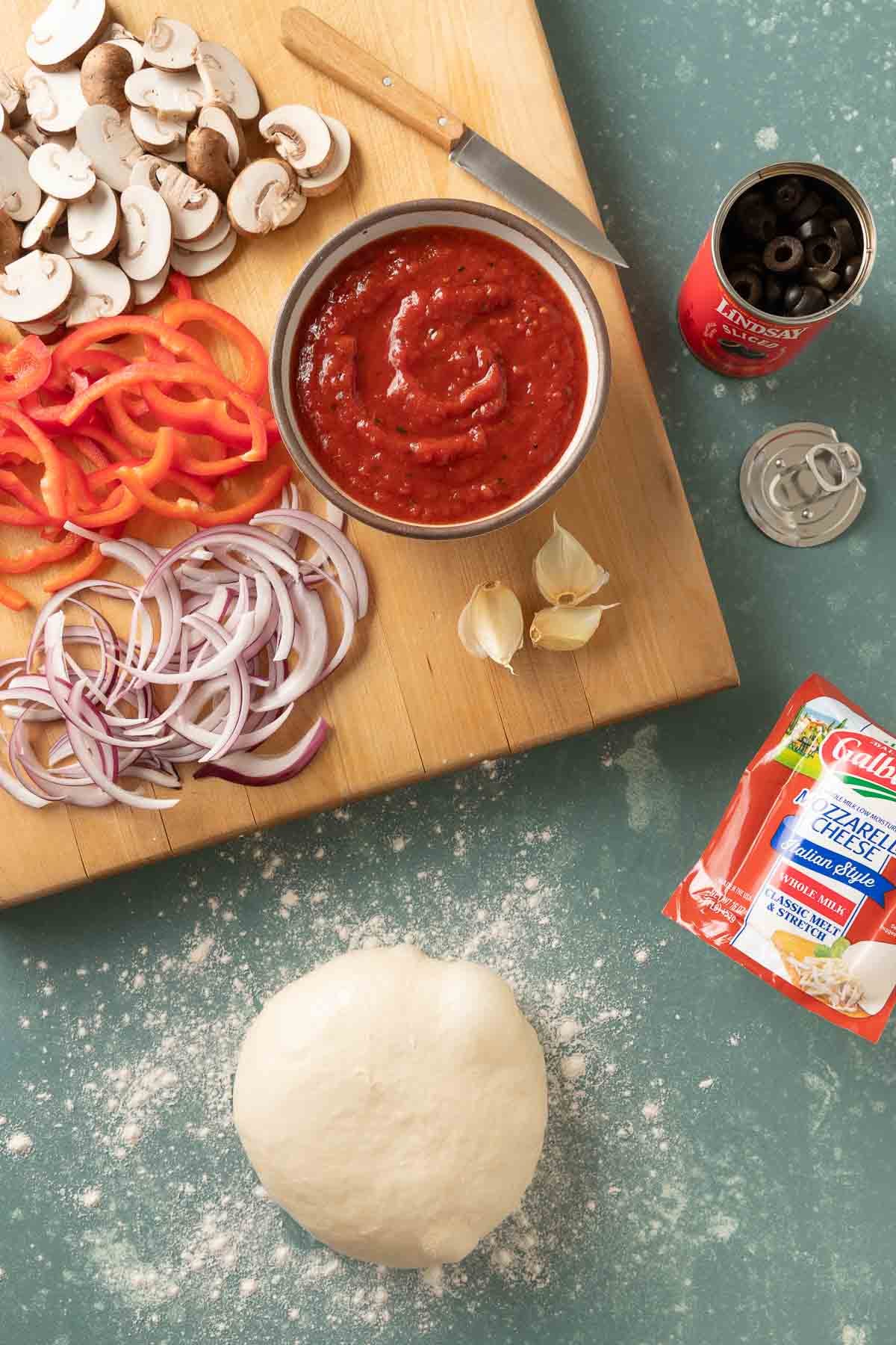 Overhead of a wooden cutting board with chopped mushrooms, bell peppers, and red onion on it with a ball of pizza dough, a small bowl of sauce and a package of cheese next to it.