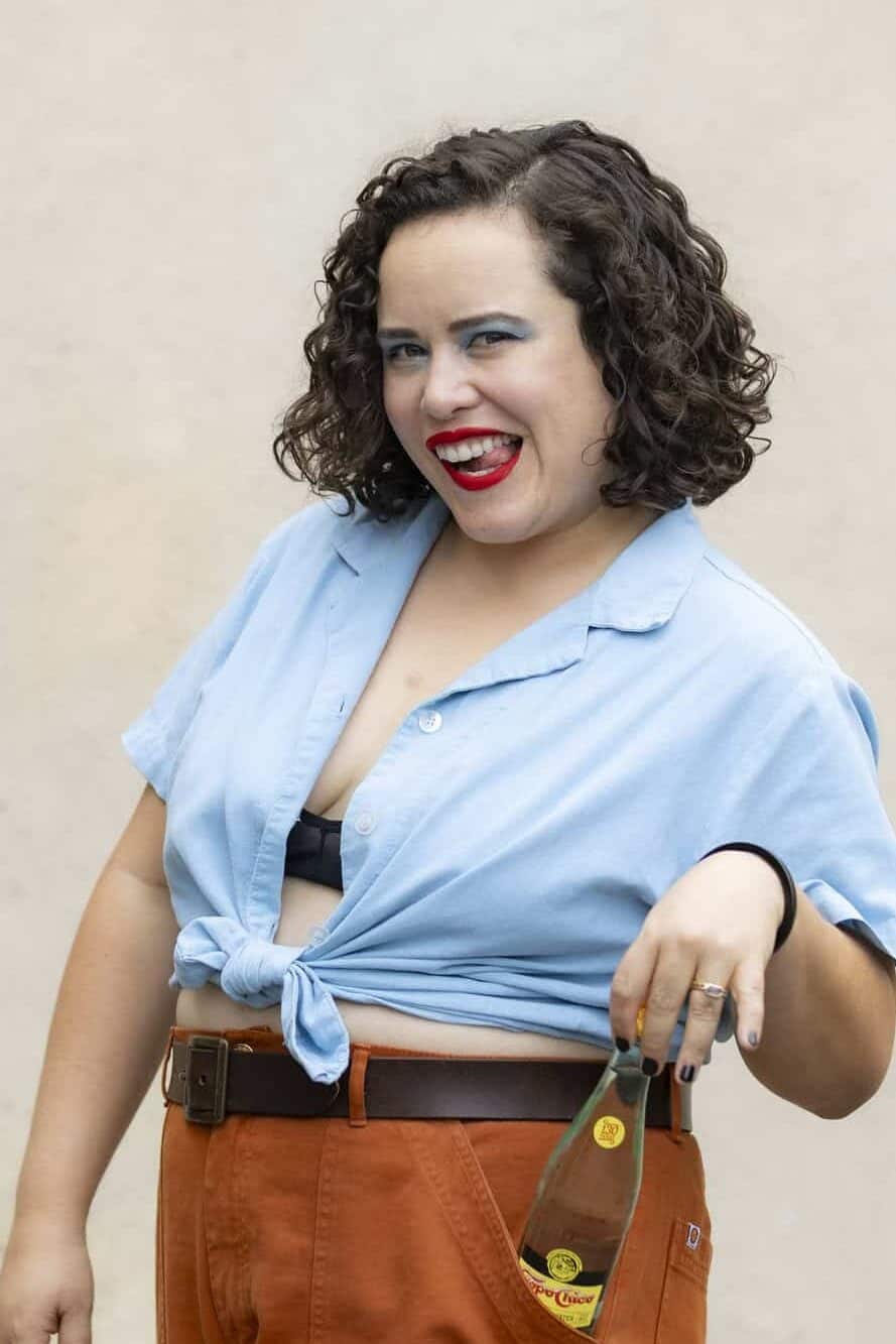 Woman with brown short curly hair smiling at the camera wearing a blue button up shirt.
