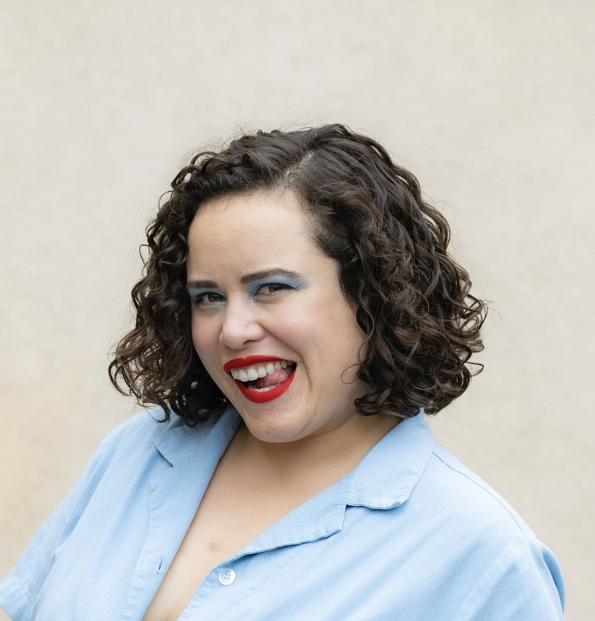 Woman with short brown curly hair, wearing red lipstick and wearing a blue button up top.