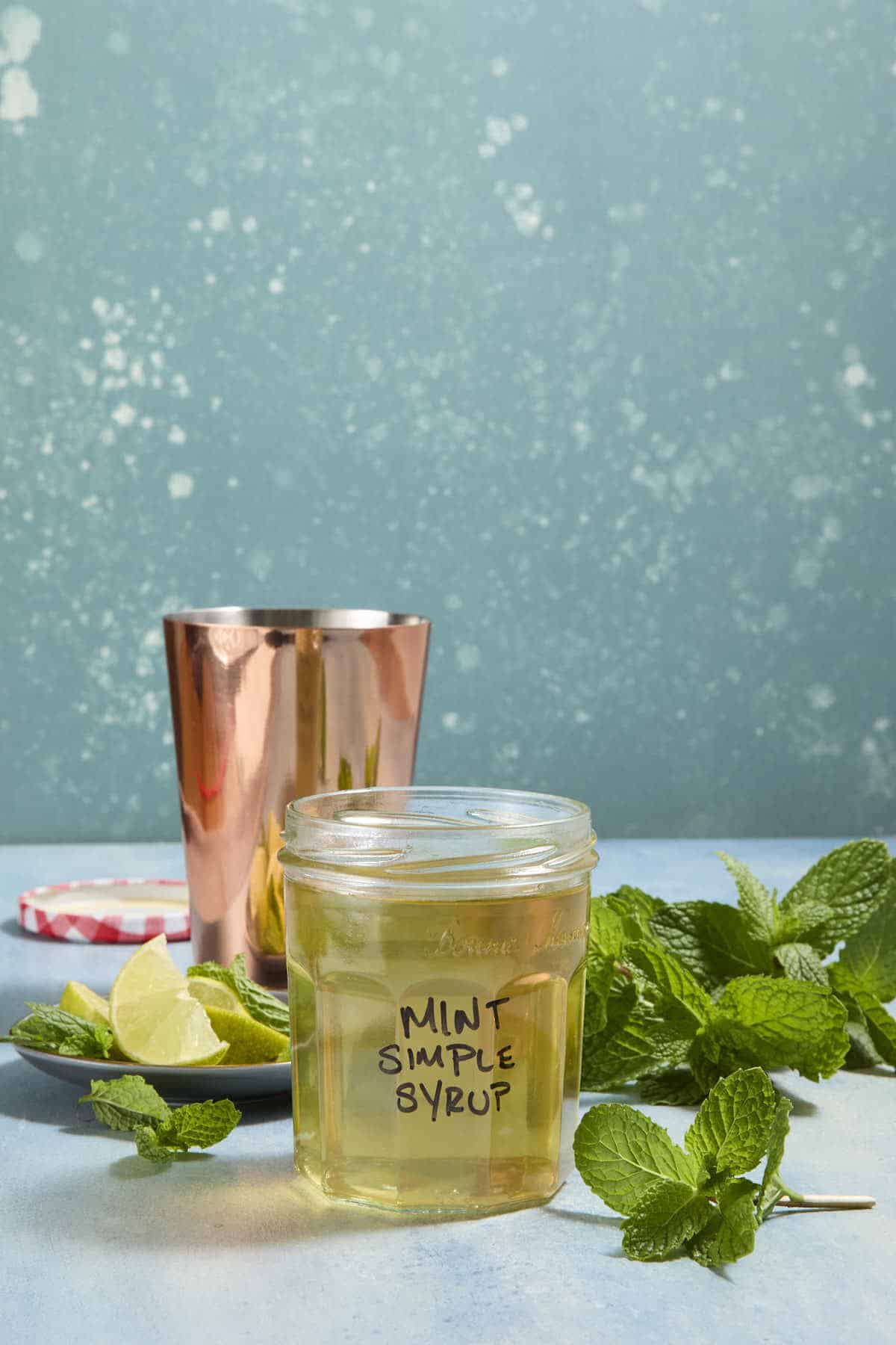 Small mason jar filled with pale yellow liquid with Mint Simple Syrup written on the front of the jar in black marker. Behind it are mint leaves and a copper drink shaker.