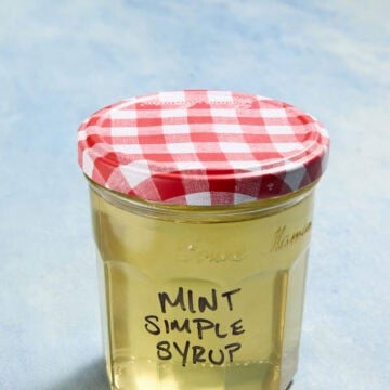 Mason jar filled with pale yellow liquid with a red and white gingham lid. Mint Simple Syrup is written in black marker on the front of the jar.