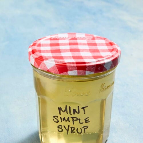 Mason jar filled with pale yellow liquid with a red and white gingham lid. Mint Simple Syrup is written in black marker on the front of the jar.