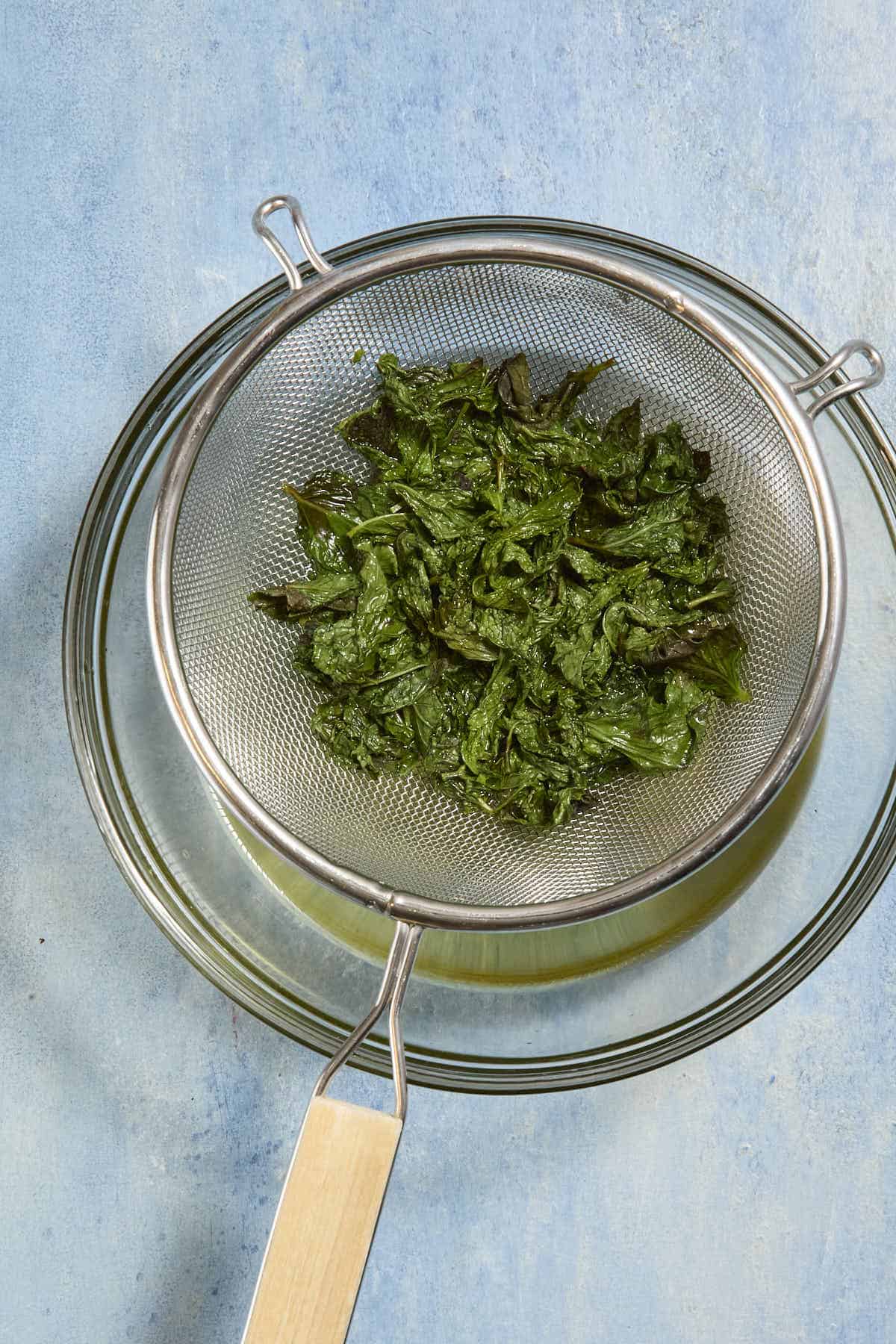 Steeped mint leaves in a fine mesh sieve draining into a glass bowl.