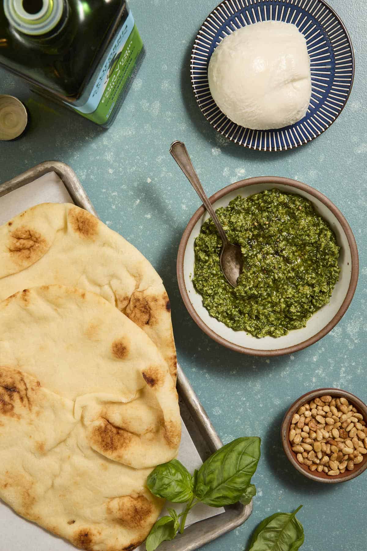 Prepared ingredients for the flatbread laid out separately, including toasted naan on a sheet tray, pesto in a bowl with a small spoon, a small bowl of pine nuts, a ball of fresh mozzarella on a blue plate and an open bottle of olive oil.