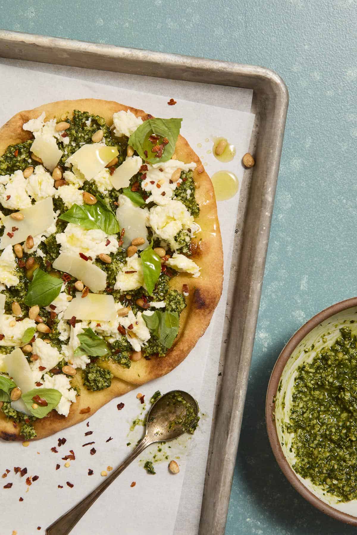 Toasted naan topped with pesto and torn pieces of fresh mozzarella and garnished with pine nuts and torn basil leaves on a sheet pan next to a spoon and bowl with more pesto.