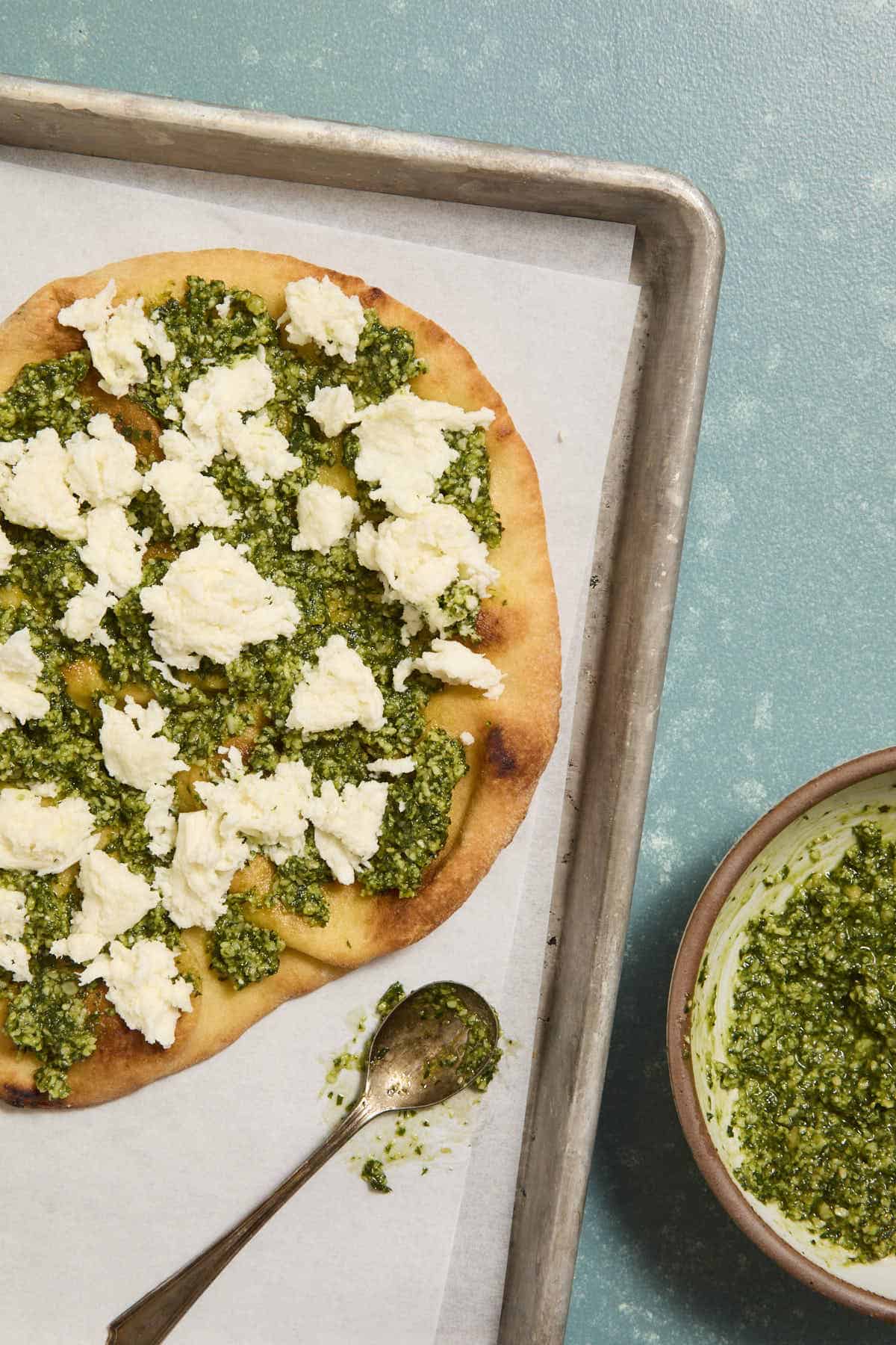 Toasted naan topped with pesto and torn pieces of fresh mozzarella on a sheet pan next to a spoon and bowl with more pesto.