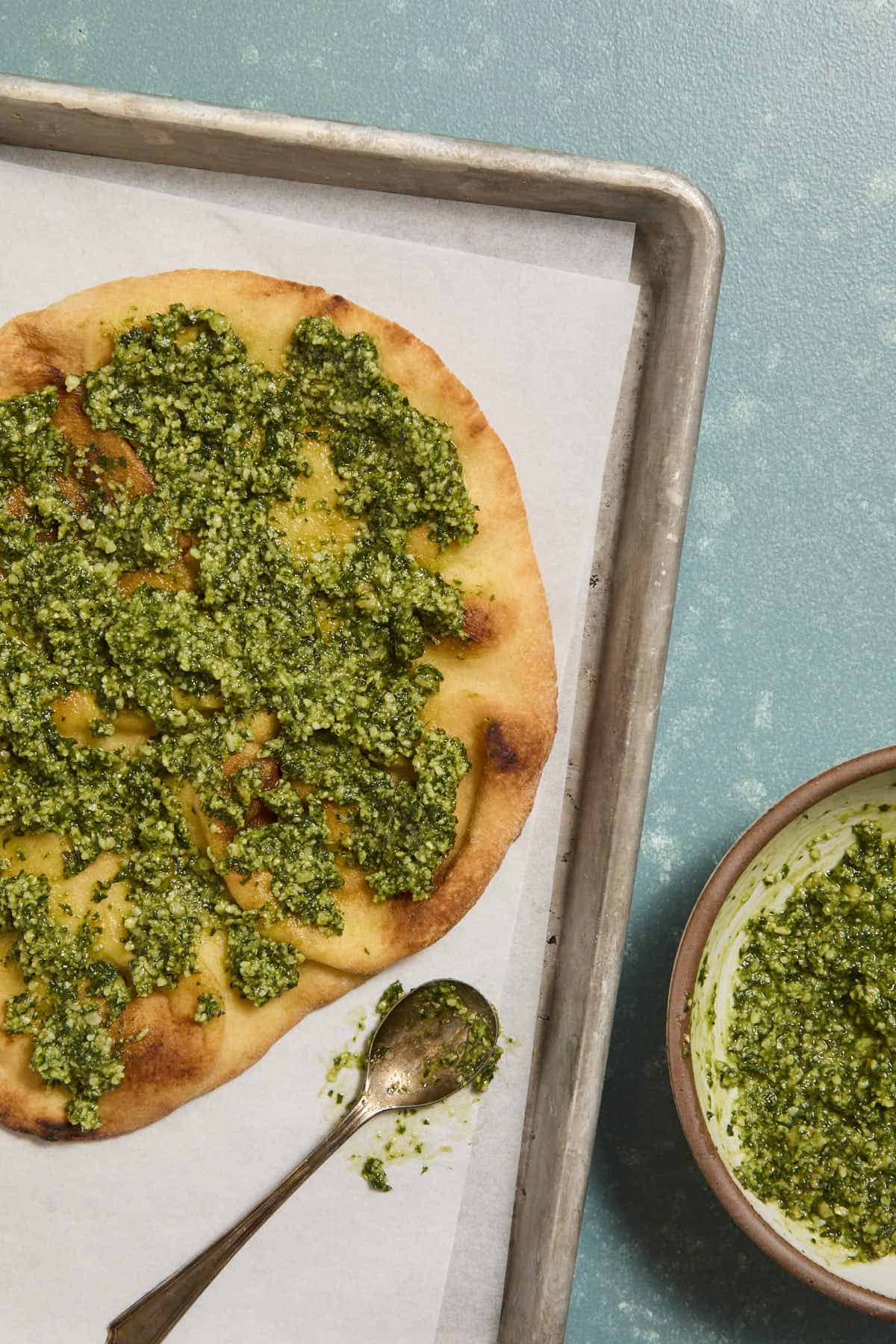 Toasted naan topped with pesto on a sheet pan next to a spoon and bowl with more pesto.